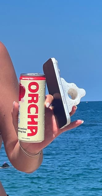 A hand holding an Apple Jasmine can and a phone in front of the ocean